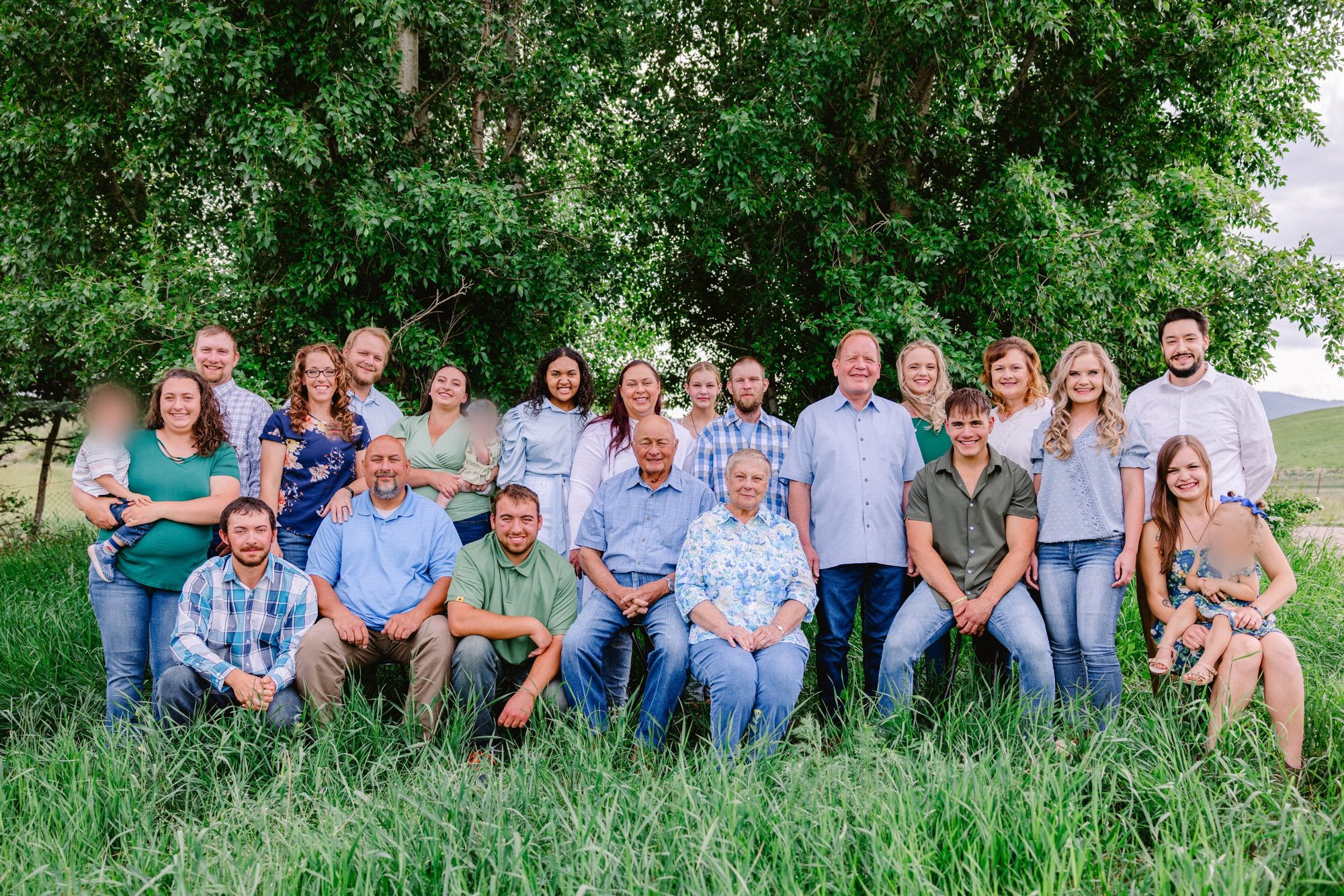 The whole extended family in 2023, gathered in tall grass under green trees, with Foalima and his wife seated in the center.